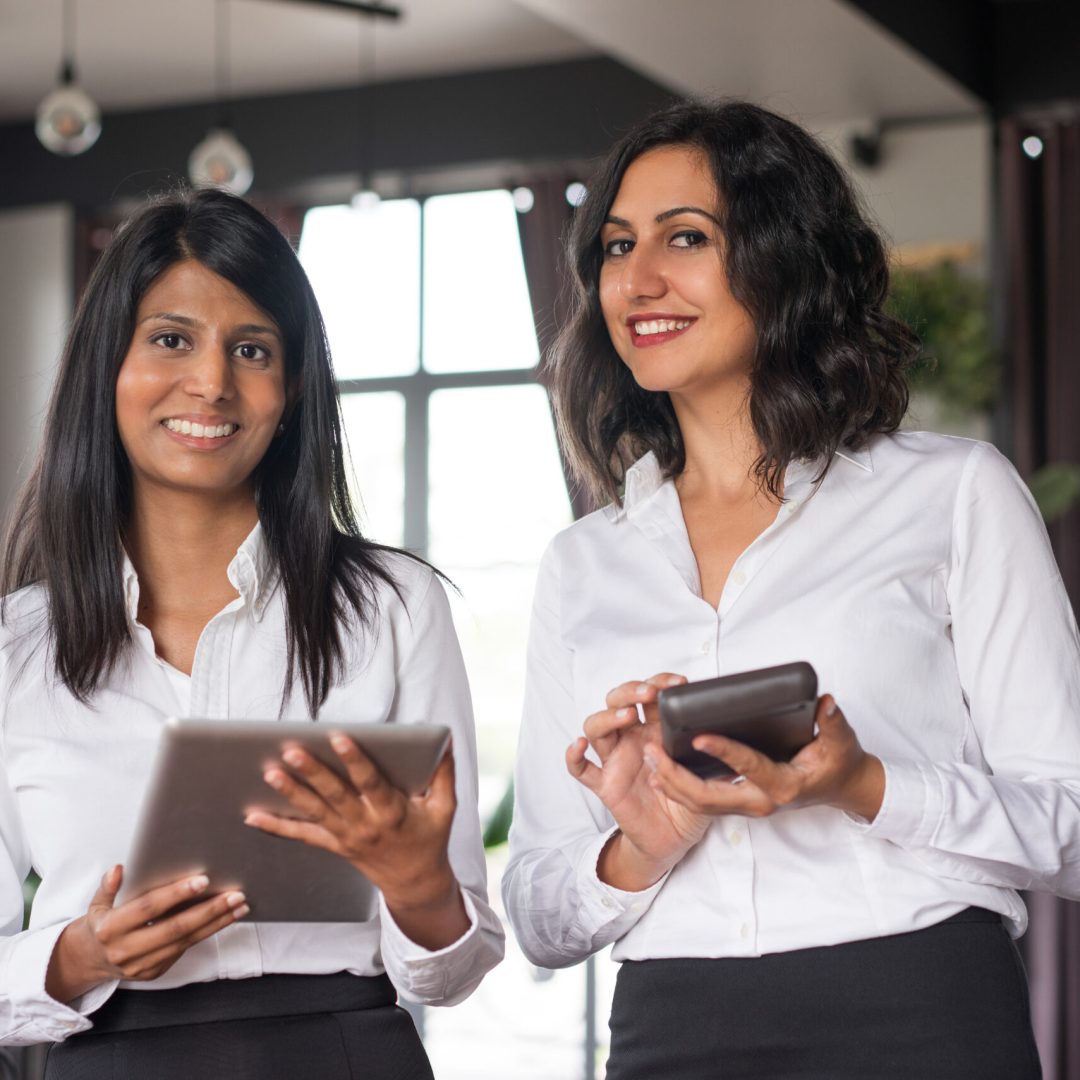 Two cheerful female colleagues using gadgets in cafe. They are standing and looking at camera with cafe interior and windows in background. Technology concept. Front view.